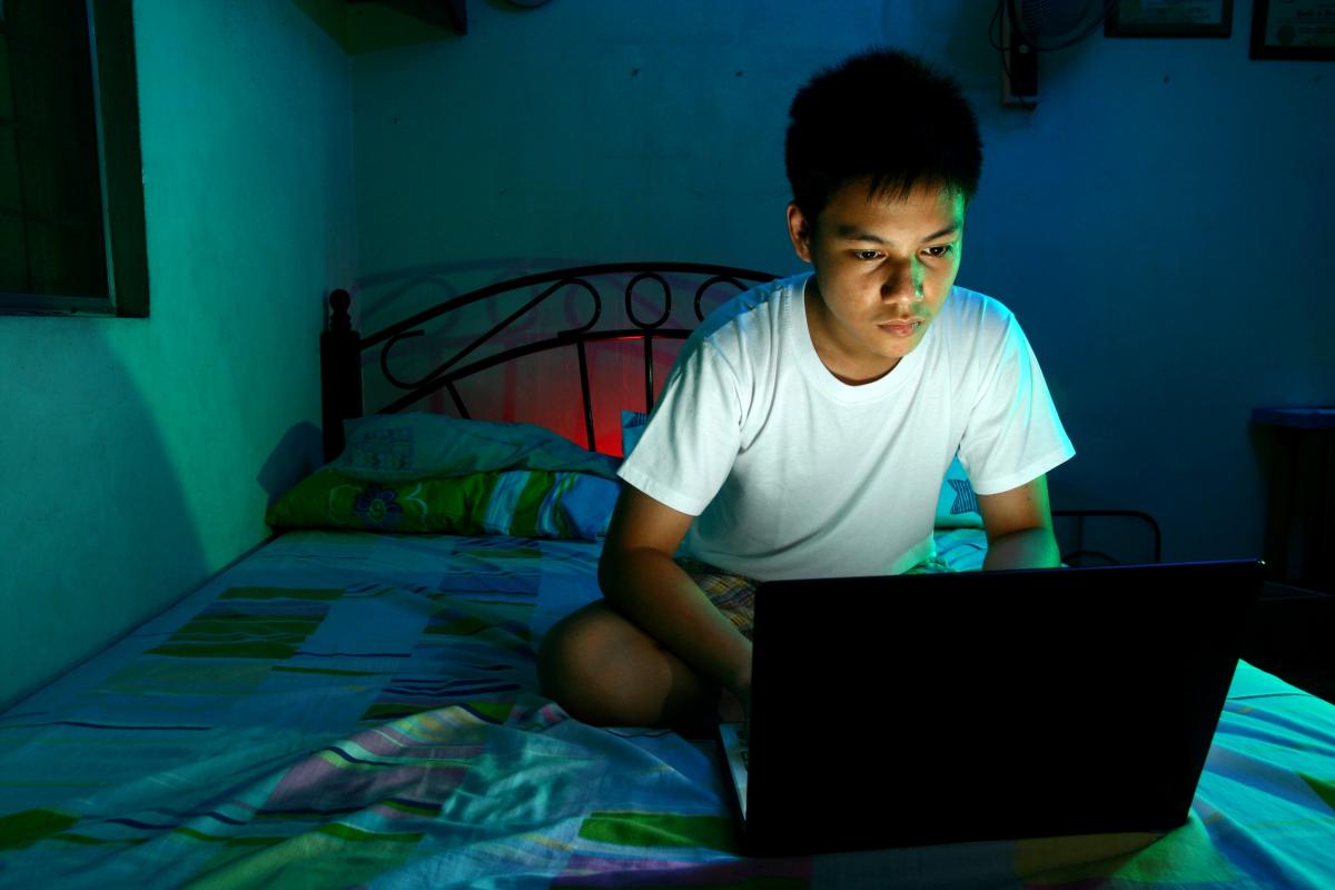 A boy sitting on his bed in the dark illuminated by his laptop screen